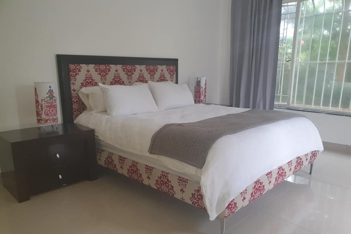 A neatly made bed with a patterned bedspread and pillows, a dark nightstand with drawers, and a large window with curtains.