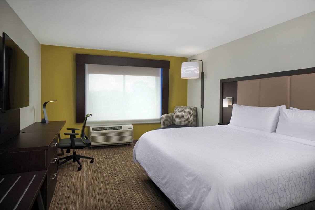 Habitación de hotel con una cama grande, un escritorio con silla, una ventana con persianas, un equipo de aire acondicionado y una lámpara.