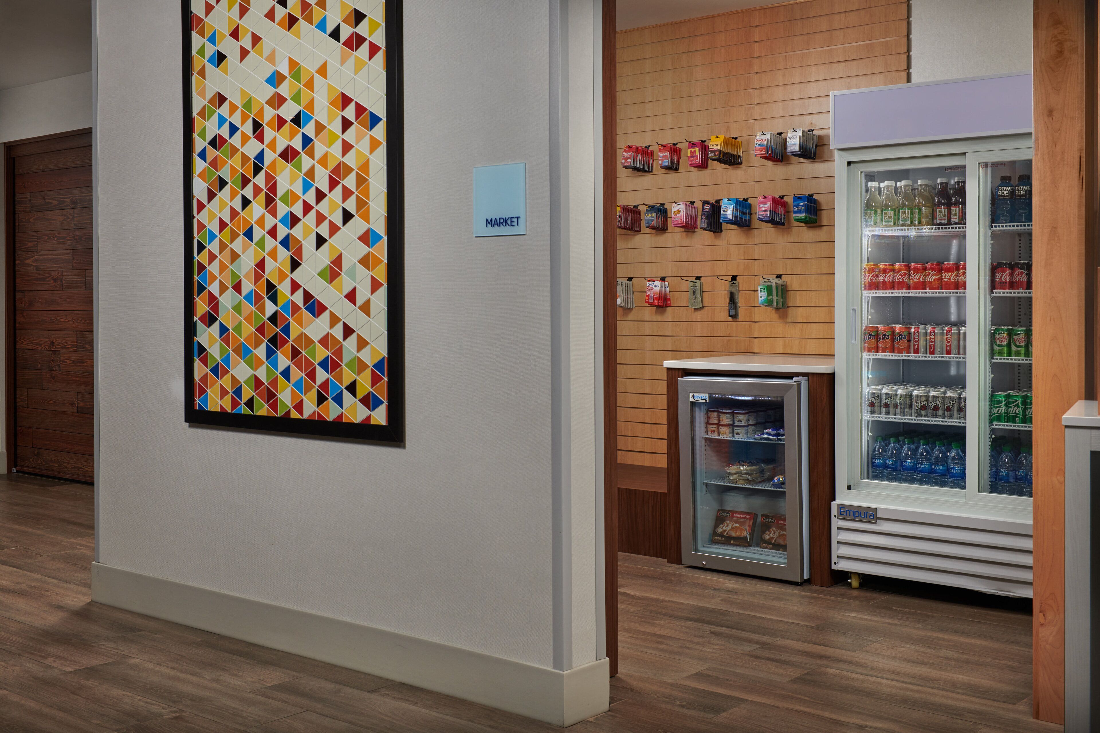 A hallway with a colorful geometric wall art, a small refrigerator with snacks, and a vending machine with beverages.