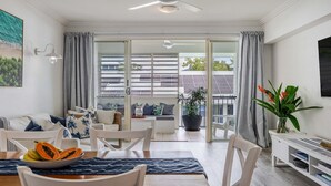 A modern living room with a dining area, large windows, and a view of solar panels. - Coastal Luxe 2BR Apartment + Spa Studio with Lagoo (PORT DOUGLAS)