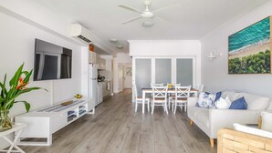 A modern living room with a flat-screen TV, a dining area with a table and chairs, a white sofa, and a ceiling fan. - Coastal Luxe 2BR Apartment + Spa Studio with Lagoo (PORT DOUGLAS)