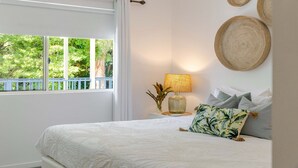 A bedroom with a bed, a nightstand with a lamp, a window with a view of greenery, and a wall with a woven item. - Coastal Luxe 2BR Apartment + Spa Studio with Lagoo (PORT DOUGLAS)