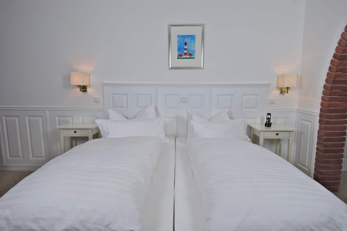 Una cama doble con ropa de cama blanca, dos mesitas de noche con lámparas, un cuadro enmarcado sobre la cama y un pilar de ladrillo a la derecha.