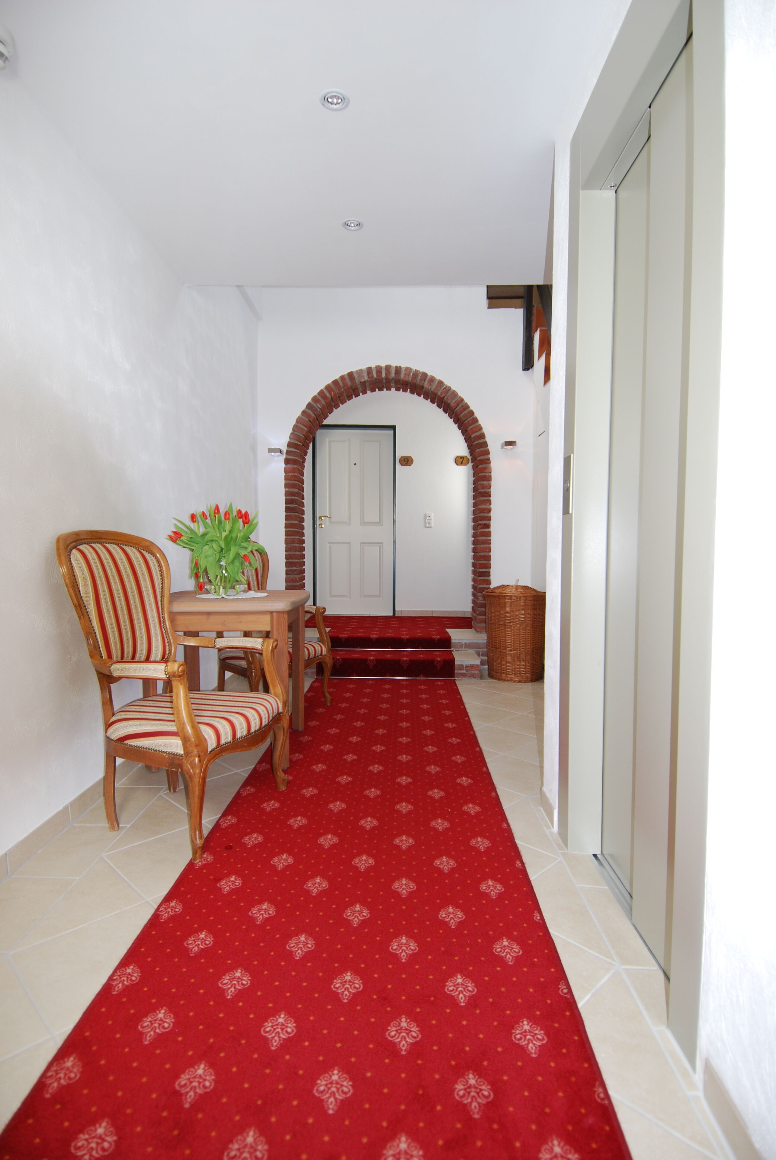 A hallway with a red carpet leading to a door, a wooden chair, a table with a vase of flowers, and a wicker basket.