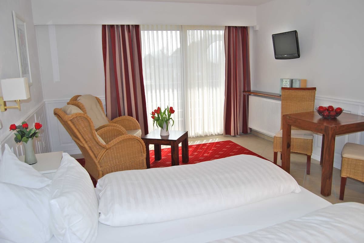 Habitación de hotel con cama, dos sillas de mimbre, una pequeña mesa con un florero, una mesa de comedor con sillas y un televisor en la pared.