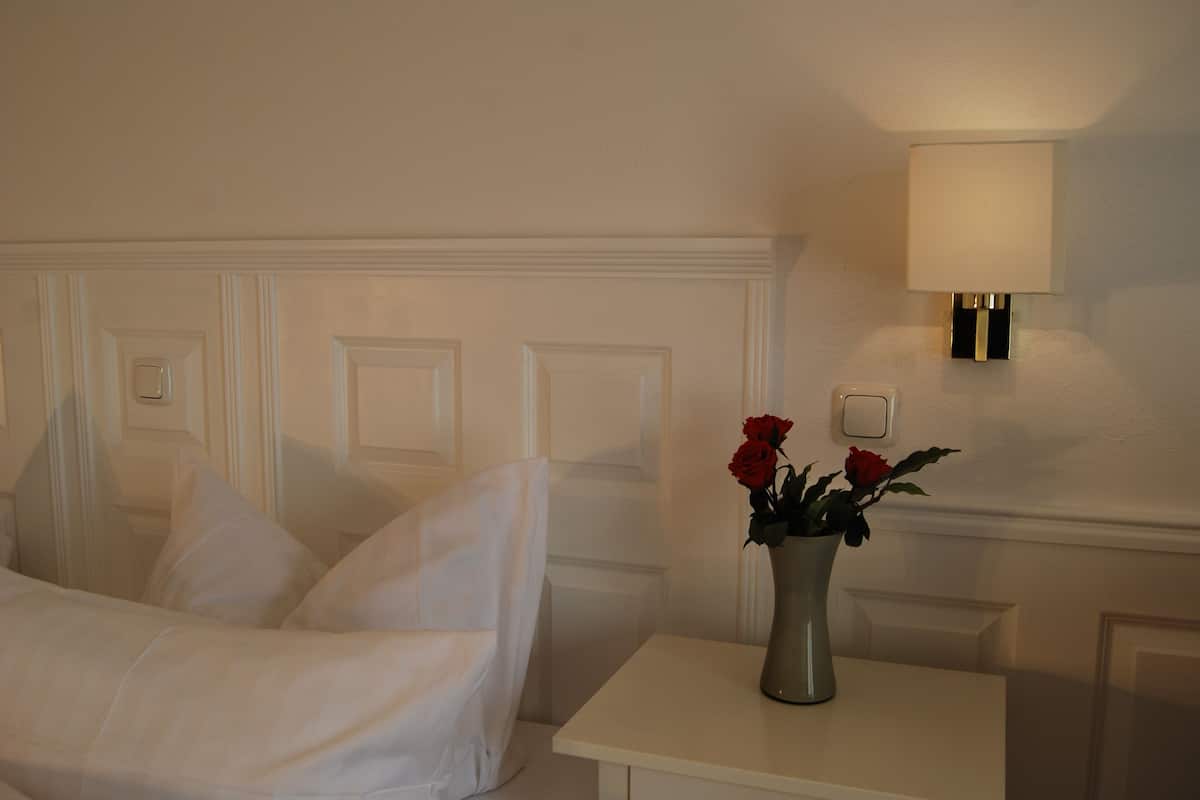 Una habitación de hotel con una cama bien tendida, una mesita de noche con un jarrón de rosas rojas y una lámpara de pared.
