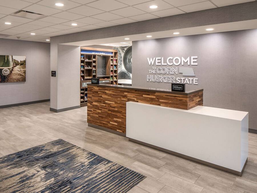 A hotel reception area with a 'WELCOME TO THE CORN HUSKER STATE' sign, a reception desk, a wooden wall, and a carpeted floor.