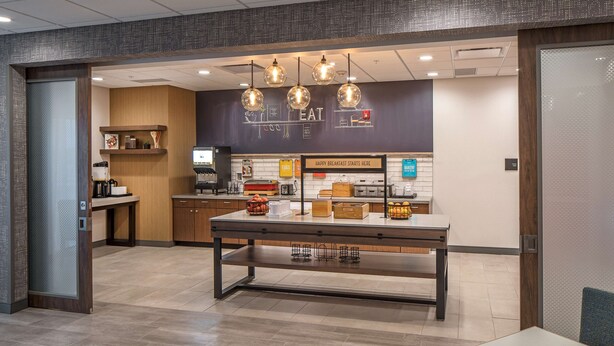 A modern kitchen area with a breakfast bar, hanging pendant lights, and a sign indicating breakfast service. - Hampton Inn Hastings (Hastings)