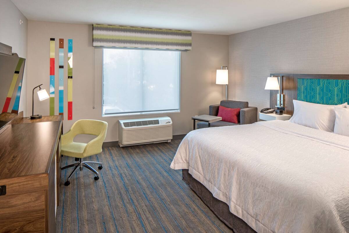 A hotel room with a large bed, a desk with a chair, a standing lamp, and a wall-mounted air conditioner.