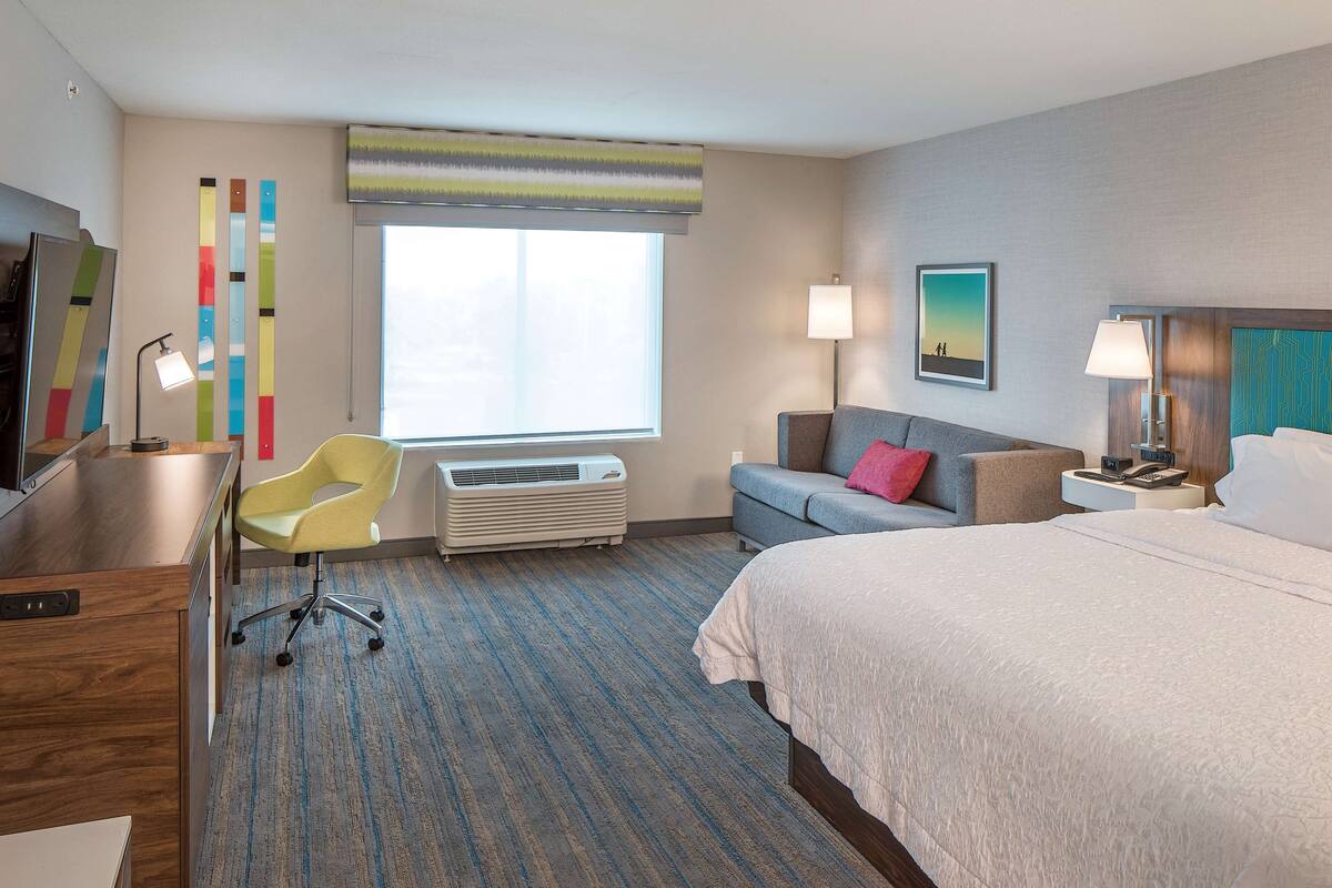 A hotel room with a bed, a desk, a chair, a sofa, a window, and a wall with vertical stripes.
