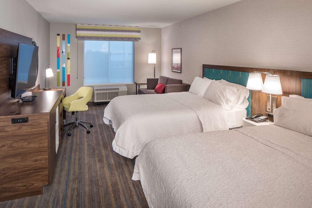 A hotel room with two beds, a desk, a chair, a TV, and a window with blinds.