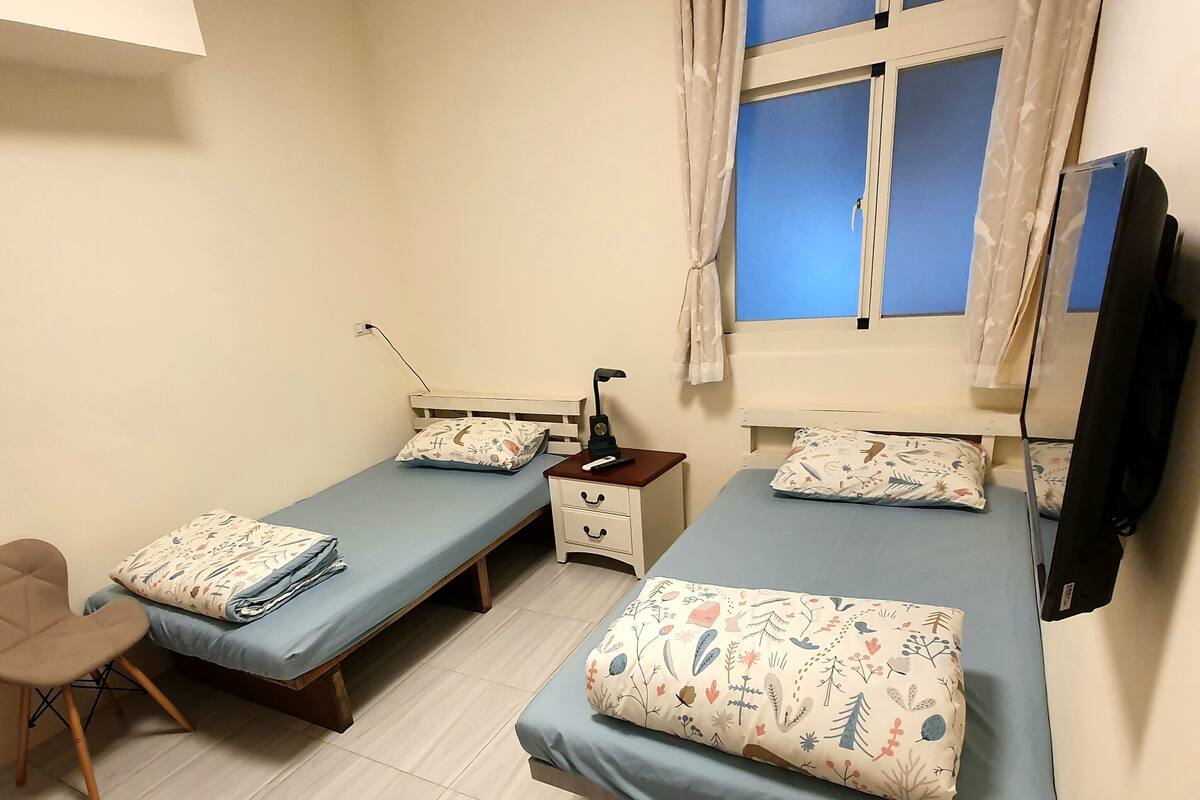 A room with two single beds, a desk, a chair, a TV, and a window with curtains.