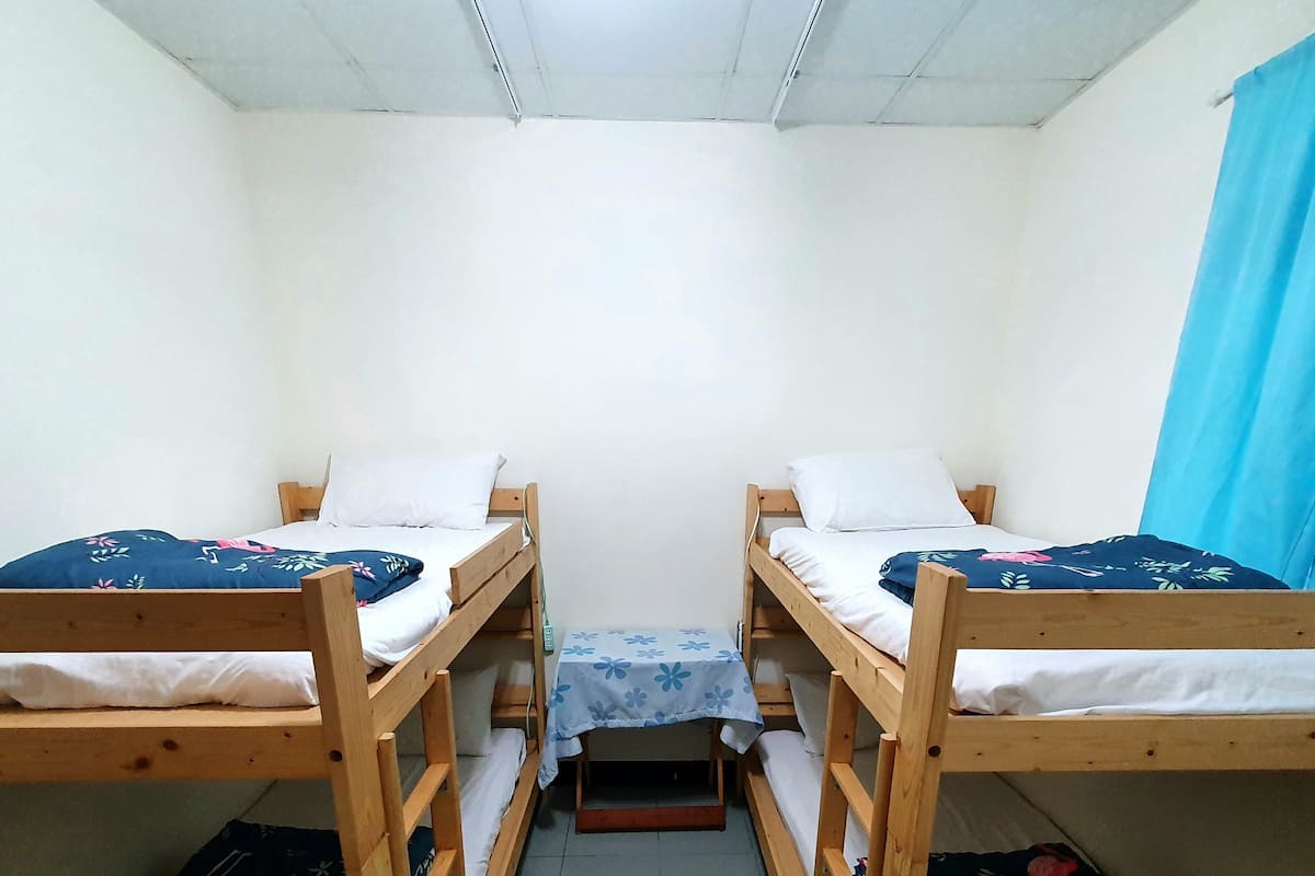 A dormitory room with two bunk beds, a small table with a floral tablecloth, and a blue curtain.