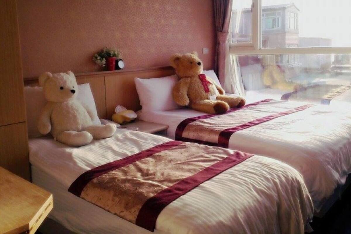 A hotel room with two beds, each with a teddy bear, a window with a view, and a bedside table with a clock.