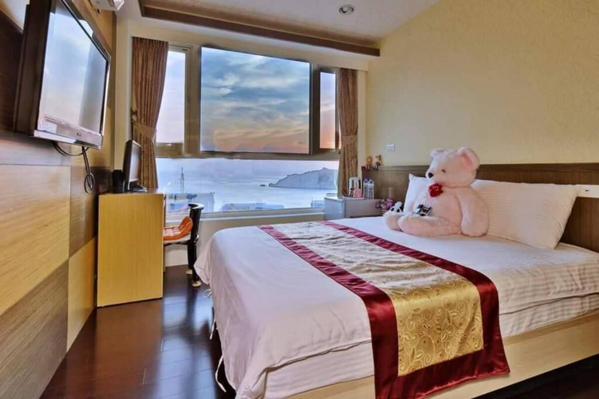 A hotel room with a bed, a desk, a television, and a large window with a view of the sea.