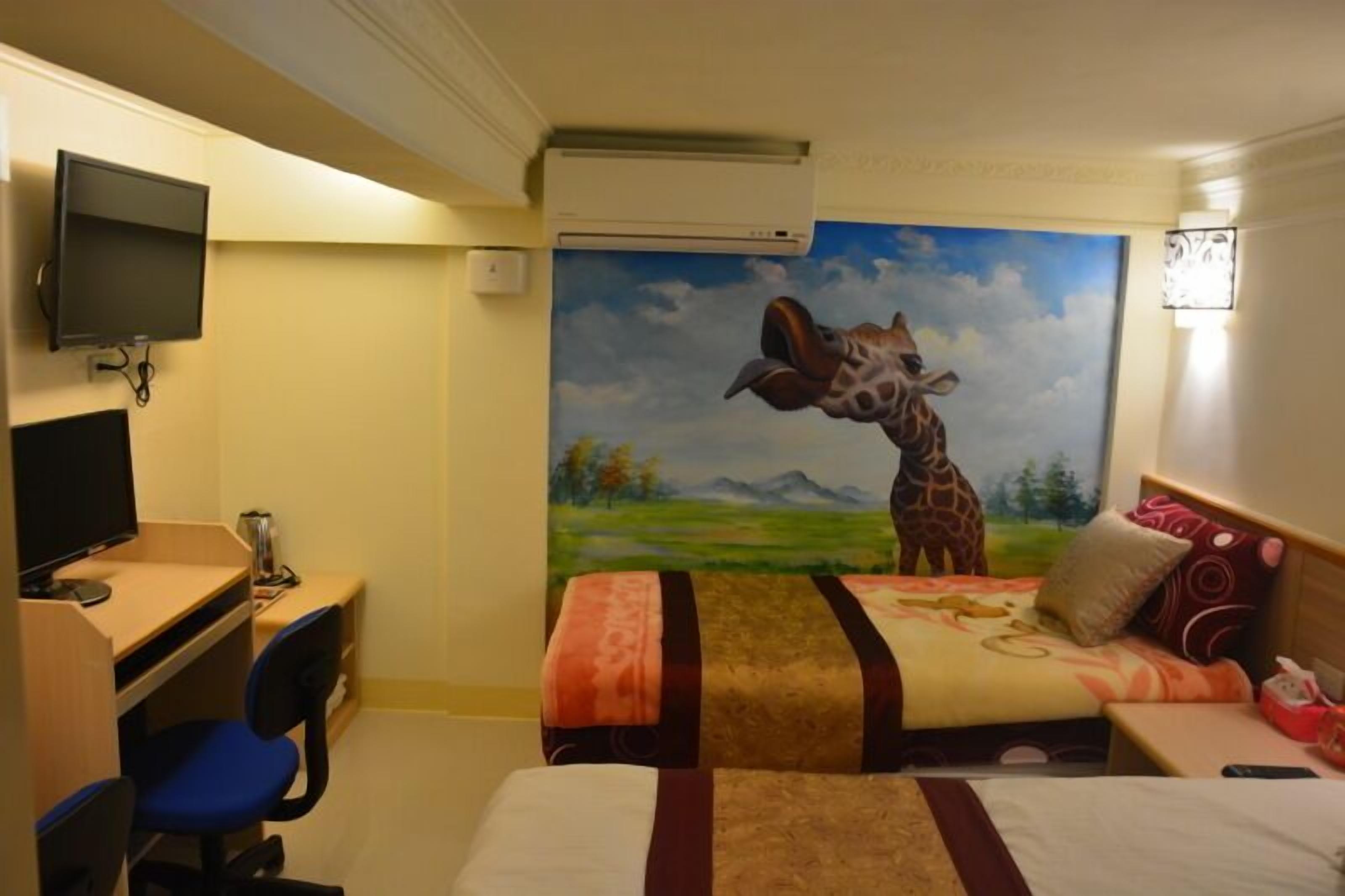 A hotel room with a desk, a bed, a TV, and a mural of a giraffe.