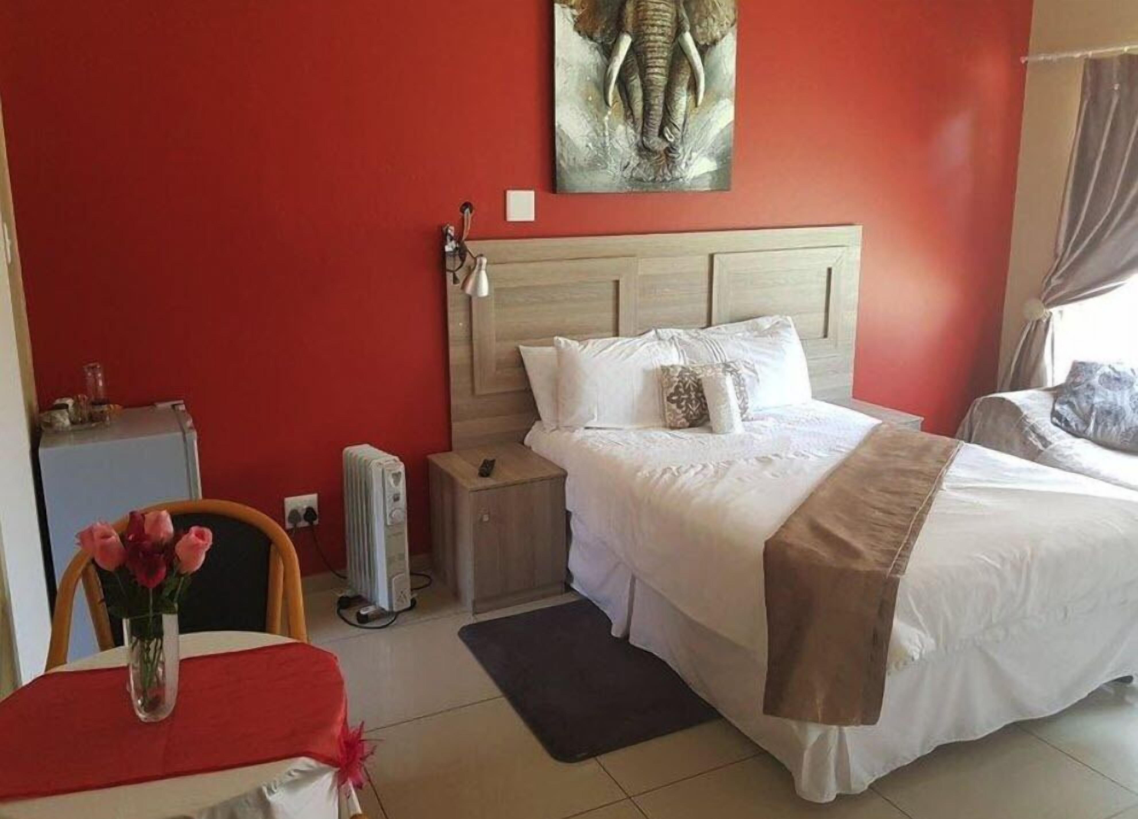 A hotel room with a red wall, a bed with white bedding, a wooden headboard, a radiator, a small table with a vase of flowers, and a painting of an elephant on the wall.