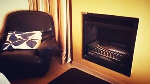 Una sala de estar con un sillón de cuero negro, un cojín estampado y una chimenea con rejilla de metal.