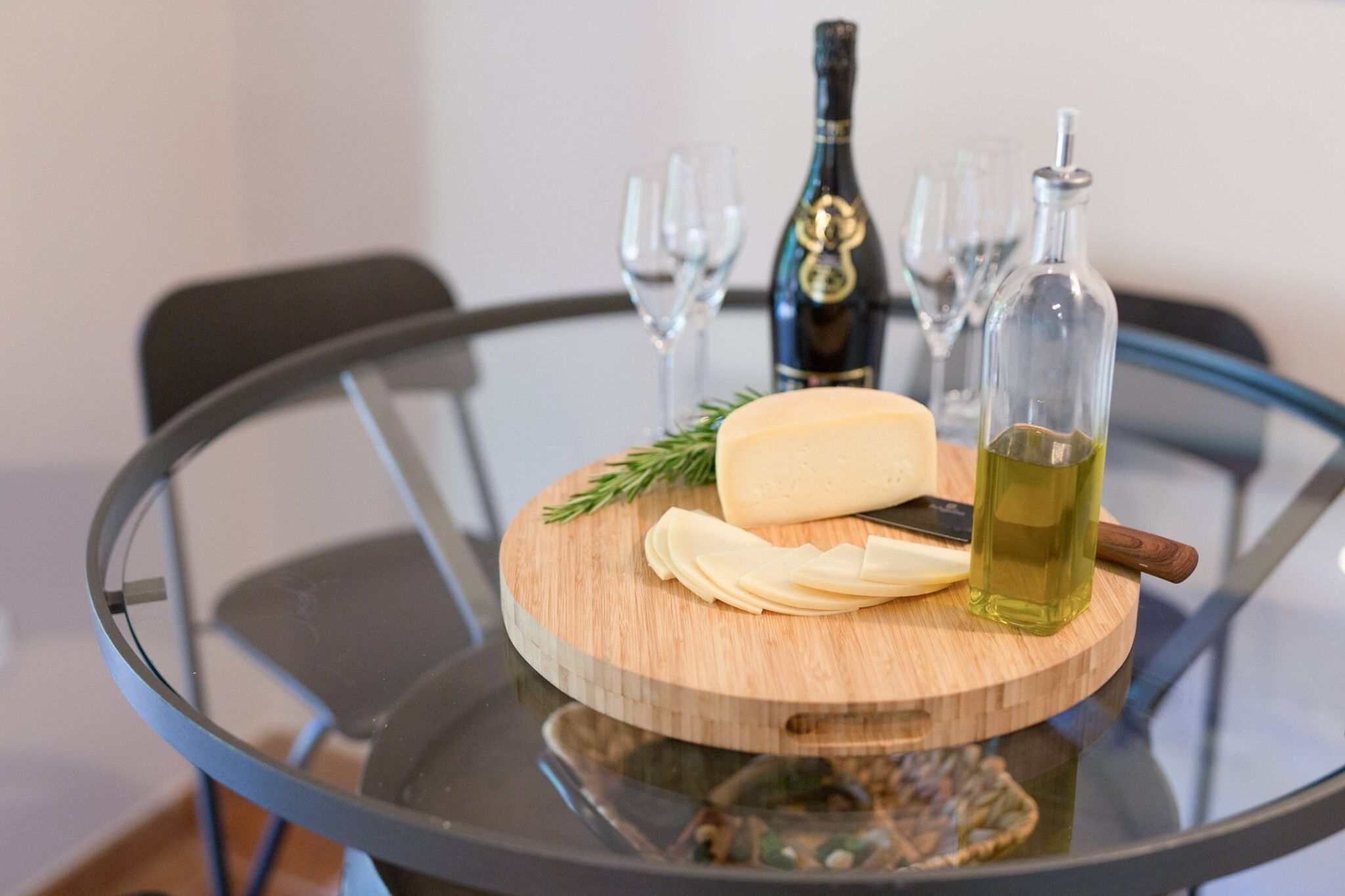 A round glass table with a wooden board holding a cheese, a bottle of olive oil, and a bottle of wine.