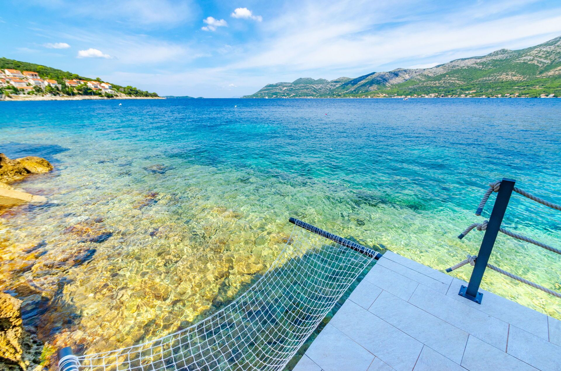 A clear view of a coastal area with a rocky shoreline, a clear blue sea, and a tiled deck with a rope barrier.