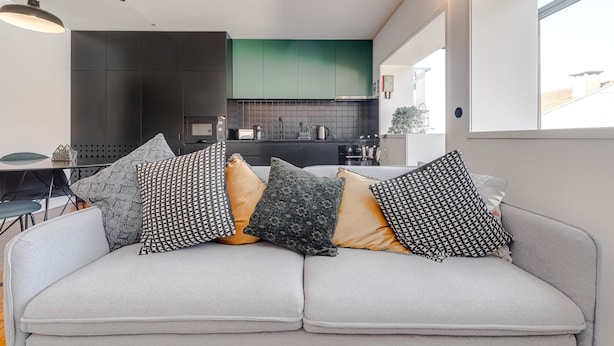 A modern living room with a grey sofa and patterned cushions, a kitchen with dark cabinets and a green backsplash, and a large window providing natural light. - Nomad's Family - 2BDR Spacious Apartment Gaia (Vila Nova de Gaia)