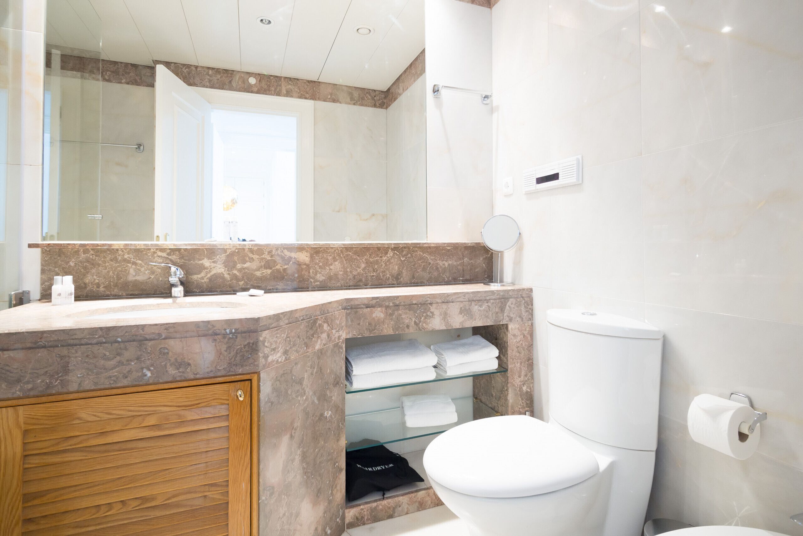 A modern bathroom with a marble vanity, a glass shelf with towels, a toilet, and a shower area.