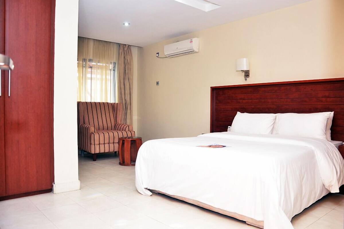A hotel room with a large bed, a wooden headboard, a striped armchair, a small wooden table, and an air conditioning unit on the wall.