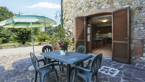 A stone building with a patio area featuring a table and chairs, an open door leading to a kitchen, and a green umbrella. - Cosy apartment in villa for 4 guests with WIFI, pool, terrace and pets allowed (Terricciola)