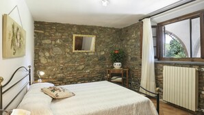 A bedroom with a stone wall, a bed with a white bedspread, a bedside lamp, a mirror, a small table with a potted plant, and a window with a view of greenery. - Cosy apartment in villa for 4 guests with WIFI, pool, terrace and pets allowed (Terricciola)