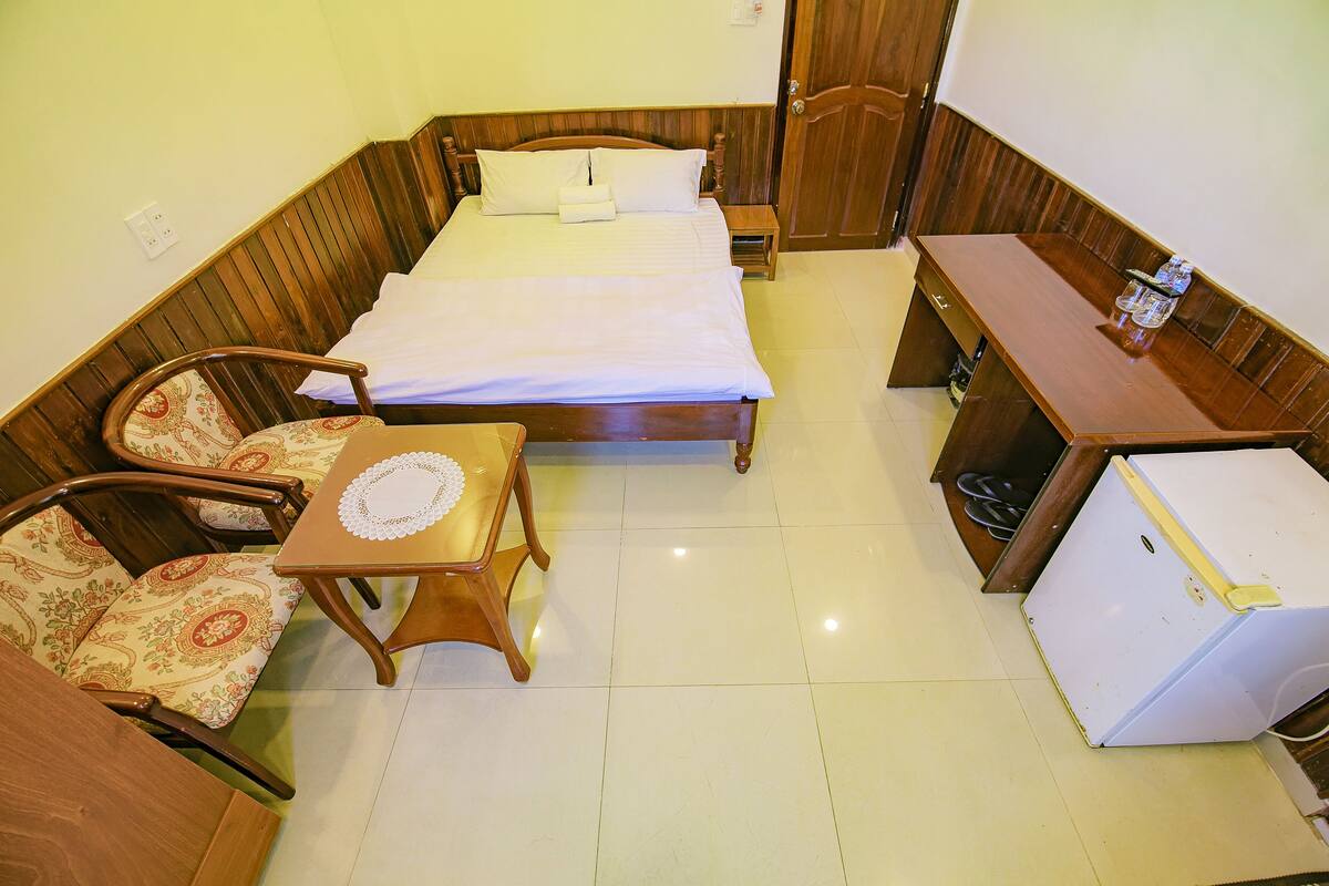 A compact room with a bed, a small table with a chair, a refrigerator, and a wooden bench.