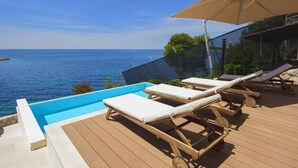 Terraza de madera con piscina, sillas de descanso y una sombrilla con vistas al mar.