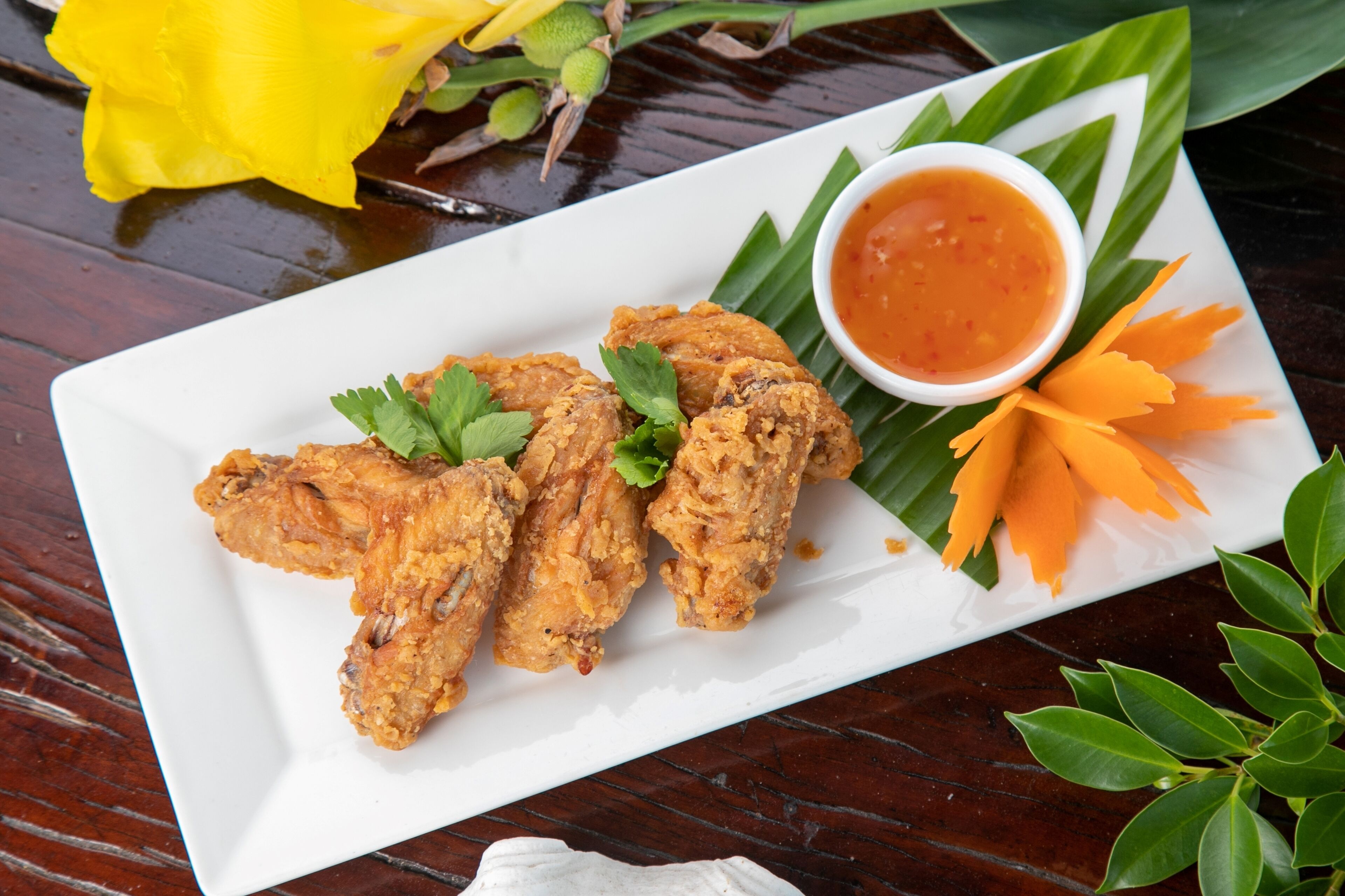 A white rectangular plate with fried chicken wings, a small bowl of sauce, and a decorative arrangement of carrot slices and green leaves.