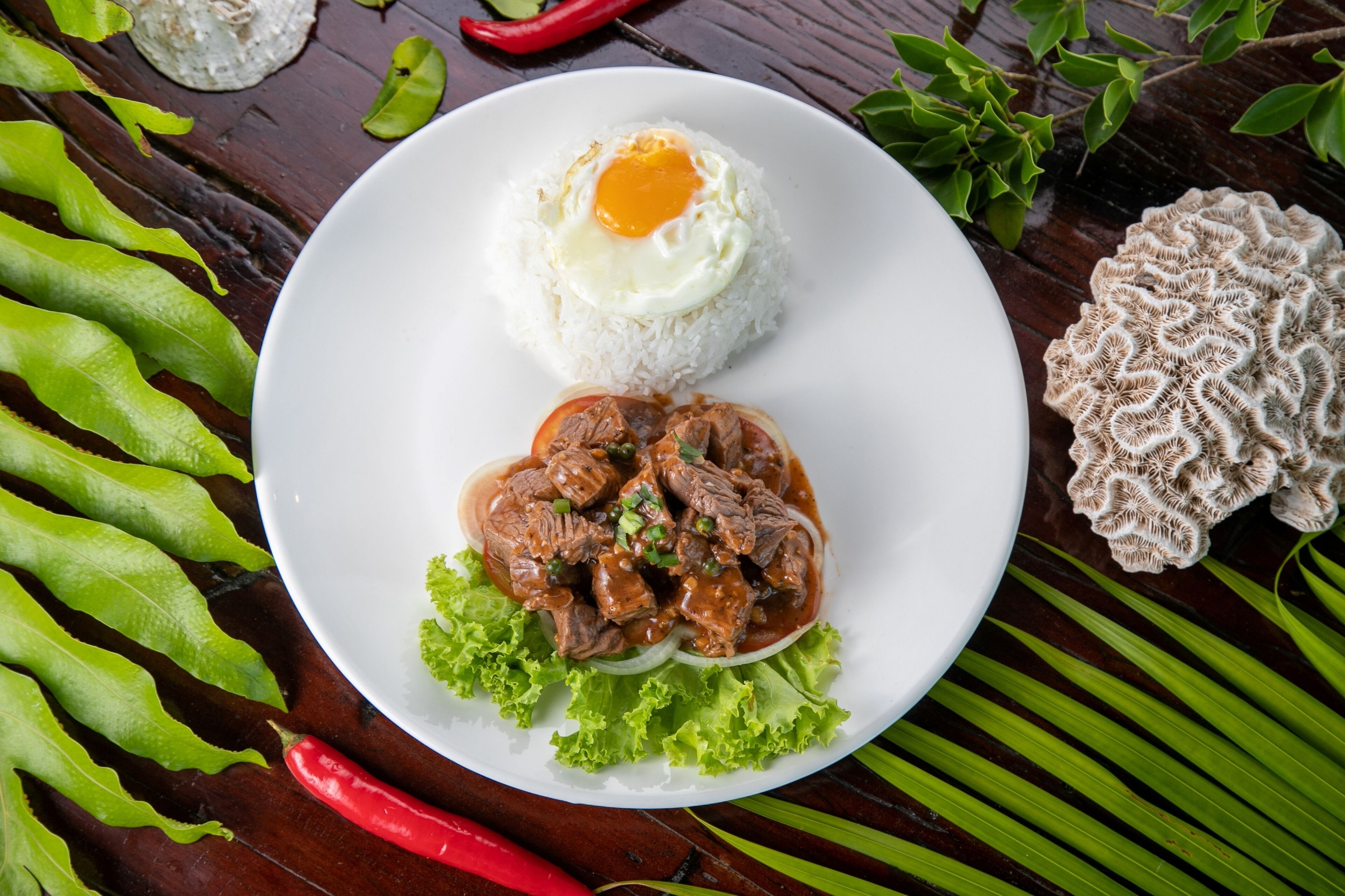Un plato de comida con arroz y un guiso de carne, rodeado de hojas verdes y un chile rojo.