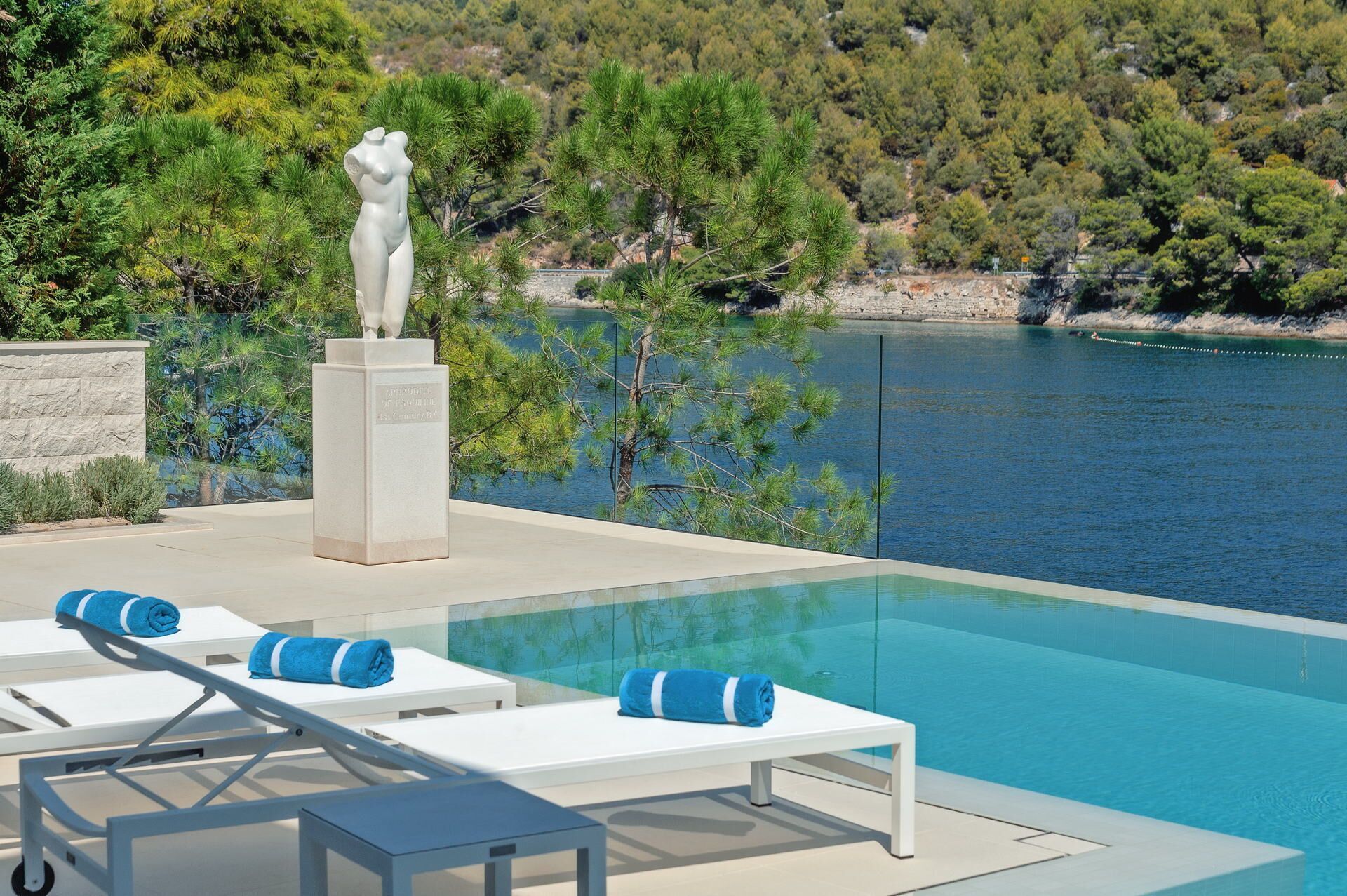 Ein Infinity-Pool mit weißen Liegestühlen und blauen Handtüchern, mit Blick auf ein Gewässer und einen bewaldeten Hügel.