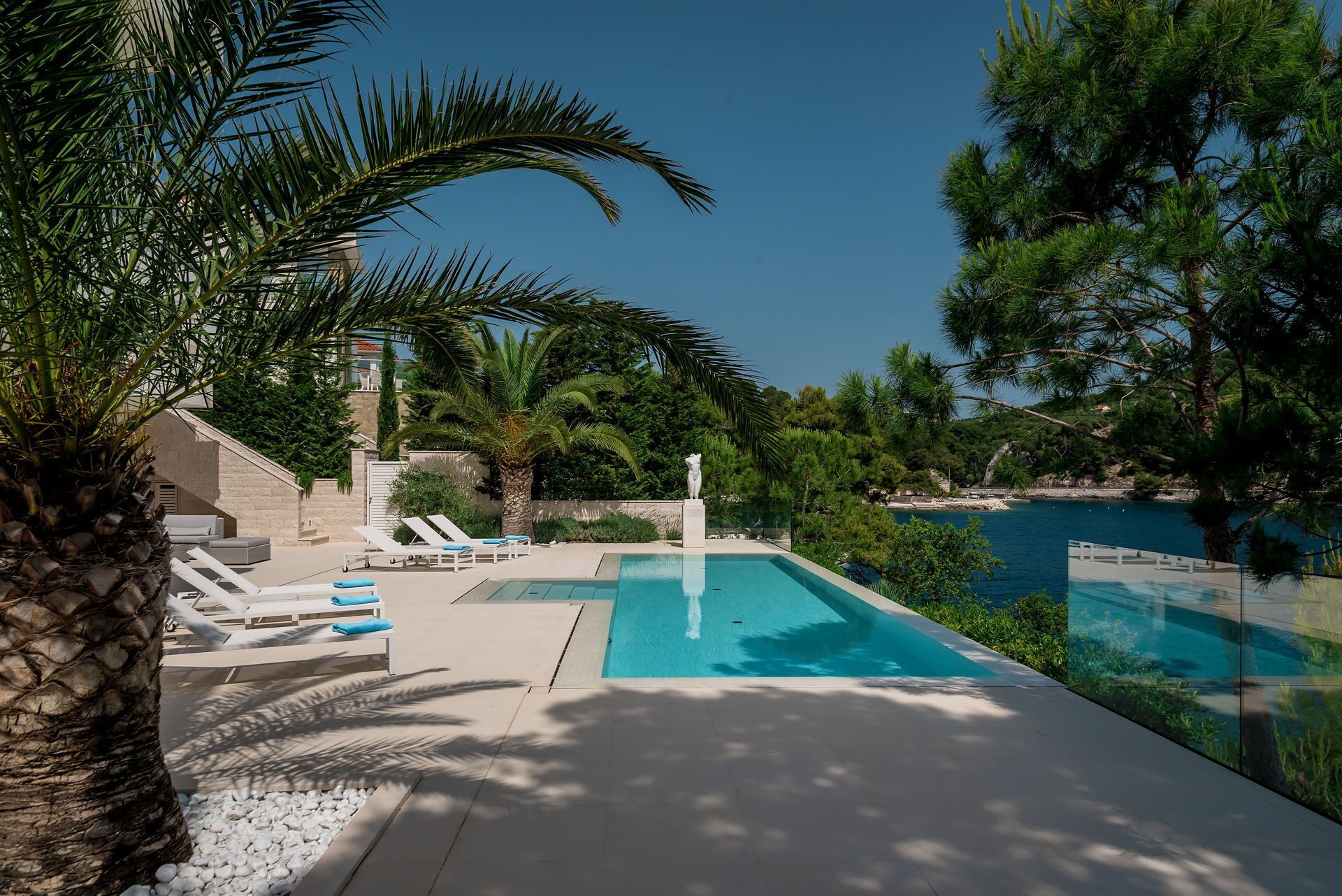 A pool area with lounge chairs and a statue, surrounded by palm trees and a clear view of the sea.