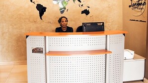 A reception desk with a world map and clocks on the wall, a sign reading "Lujuria COTTAGE," and a small desk with a printer. - Lantano Cottage (Lagos)