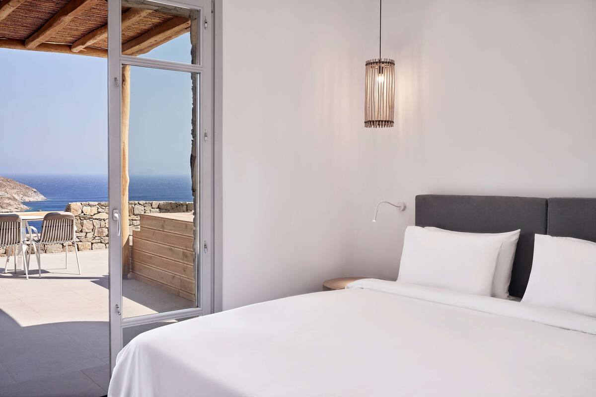 A bedroom with a bed, a nightstand, and a hanging light fixture. There is a glass door leading to an outdoor area with a view of the sea.