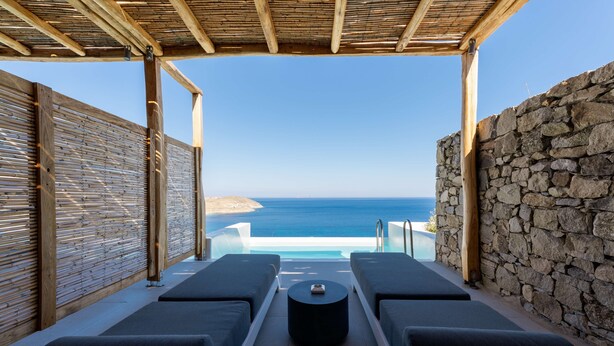 A sunlit outdoor lounge area with two lounge chairs and a small table, overlooking a clear blue sea and a rocky coastline. - Radisson Blu Euphoria Resort, Mykonos (Mykonos)