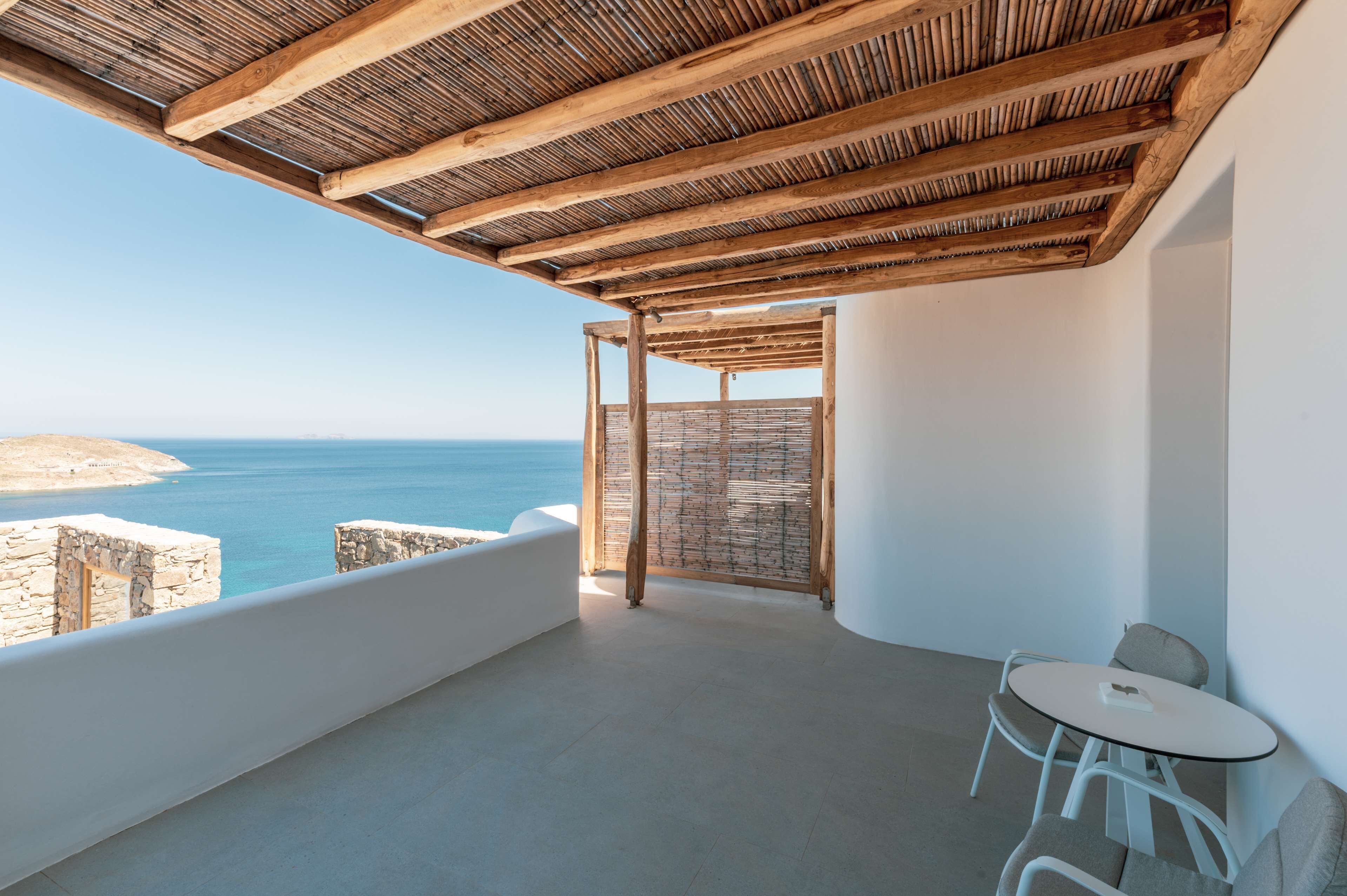 A balcony with a wooden ceiling, a table, and chairs, overlooking a coastal view.