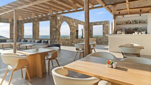 An outdoor seating area with stone arches, wooden tables, and chairs, overlooking a scenic view. - Radisson Blu Euphoria Resort, Mykonos (Mykonos)