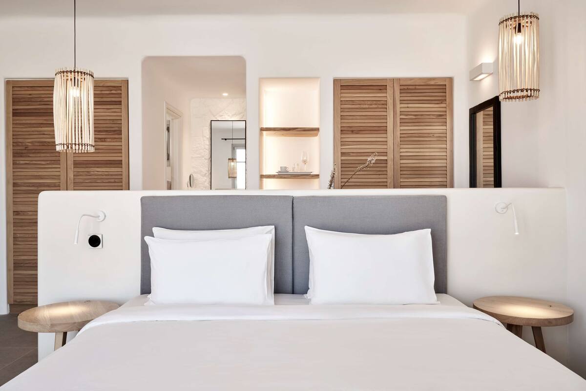 A modern hotel room with a large bed, two bedside tables, a mirror, and a wooden wardrobe.