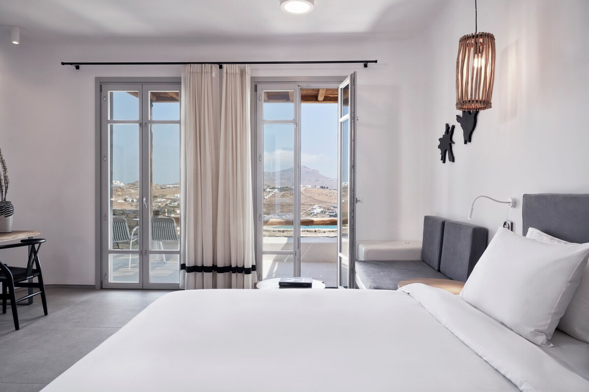 A modern hotel room with a large bed, a desk, and a balcony with a view of the landscape.