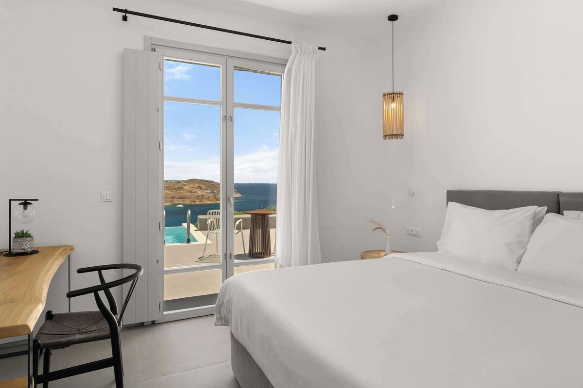 A bedroom with a bed, a desk, a chair, and a view of the sea through a glass door.