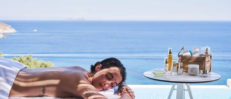 A person relaxing by a pool with a view of the sea, a table with skincare products, and a towel.
