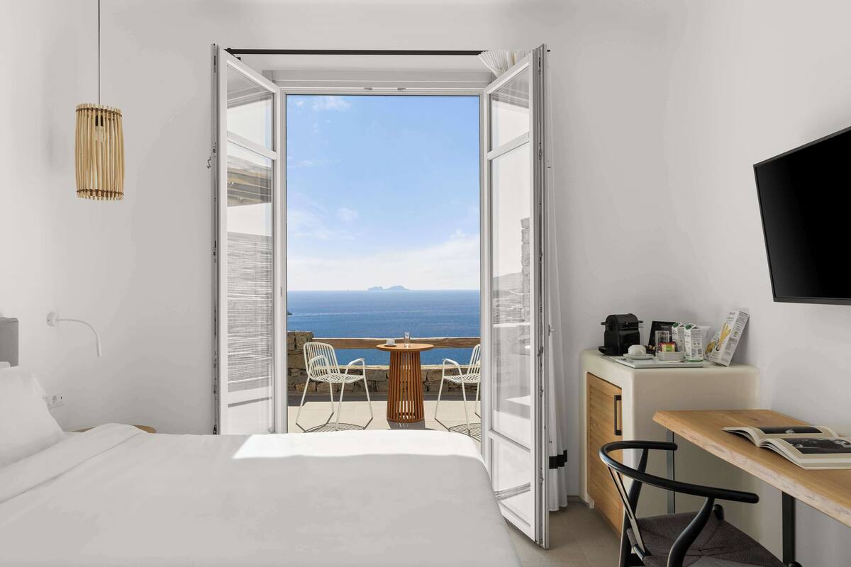 A hotel room with a bed, a table with chairs, a TV, and a balcony with a view of the sea.
