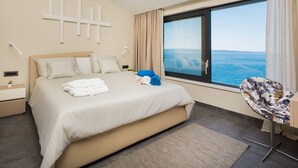 Habitación de hotel con una cama grande, vista al mar, una silla y una lámpara de pared.