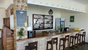 A bar area with a stone counter, multiple bar stools, a refrigerator, and a shelf with various bottles. - Jazzybeach Hotel (Nosy Be)