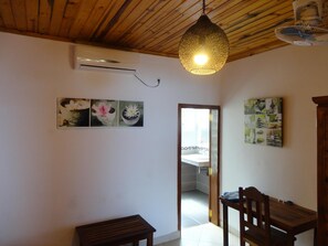 A room with a wooden ceiling, a hanging light fixture, an air conditioning unit, a dining table with chairs, and a door leading to another room. - Jazzybeach Hotel (Nosy Be)