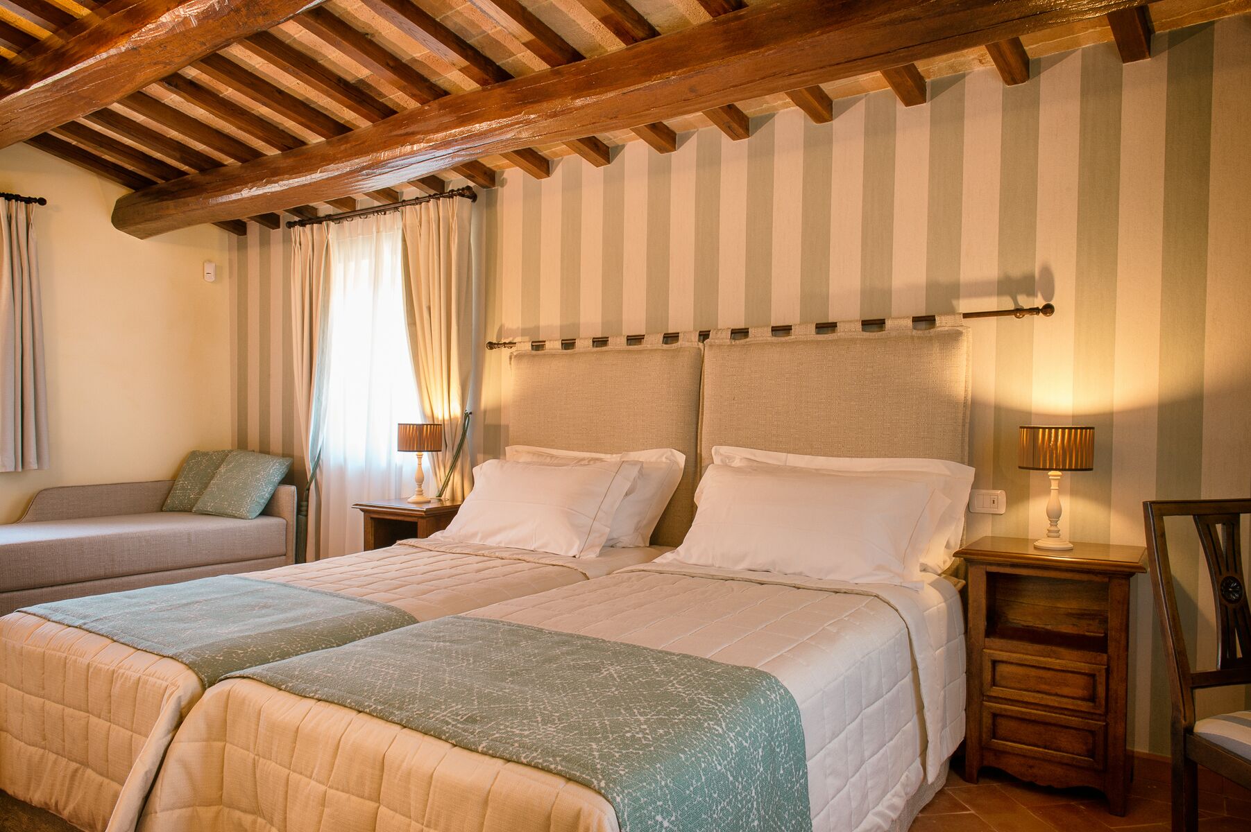 A double bed with a headboard, a bedside table with a lamp, and a sofa in a room with striped wallpaper and wooden beams.