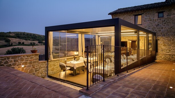 A modern glass-enclosed dining area with a stone building in the background. - Borgo Sant'Apollinare (Marsciano)